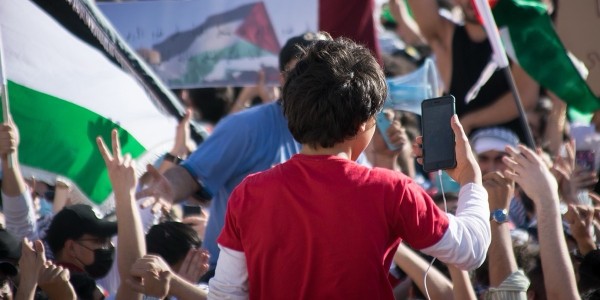 A child in a red shirt holding up a smartphone surrounded by a crowd with raised hands and Palestinian flags.