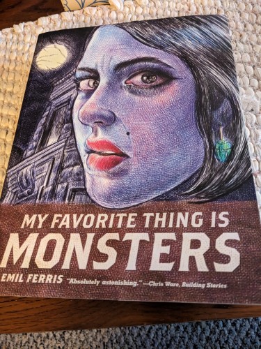 A thick, soft cover copy of My Favorite Thing is Monsters.  A three-quarter profile of a white woman with dark hair, a green earring, a mole, and red lips. The art style is sketchy and features lots of cross-hatching.  A full moon and a sliver of a Chicago townhouse are in the background.