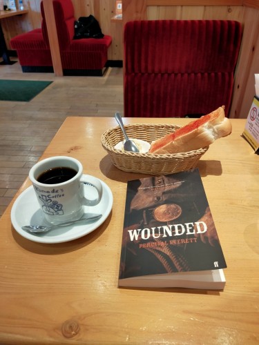 Photo is of a light brown wooden cafe table  The paperback book is old timey brownish focusing on a coppery belt buckle of a cowboy. Behind the book is a small narrow wicker basket perpendicular to the book. It contains a tiny silver spoon in a tiny white bowl containing a dollop of yellow and white scrambled eggs and a long protruding thick rectangular slice of toast with red strawberry jam on it. To the right is a white mug and saucer of black coffee. It has the Komeda Coffee logo of a man in a cowboy hat drinking coffee. There is a tiny silver spoon in front of it, handle pointing left