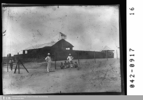 Black and white photograph showing a large building with an old German flag on top. It is surrounded by a barbed wire fence. Outside the fence there is a group of 5 apparently African persons sitting on the floor. Two white men in white uniforms and helmets stand next to them. One African soldier with a rifle is standing behind the fence apparently guarding the building.