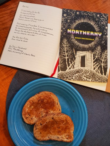 Poetry collection Northerny by Dawn Macdonald (University of Alberta Press), with its striking black and white images on the cover, sits on a notebook in which is transcribed the poem "Aperture", along with toast and marmalade on a blue plate