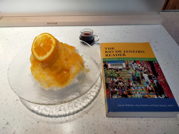 Photo is of a marble counter on which is the paperback book where the top title is black text in orange, the subtitle text is white in green. The bottom blue has white text of editor's names. In between is a colorful photo of Brazilians of various ethnicities sitting along a colorful outside stairs. To the left is orange shaved ice in a glass bold. An orange slice is on top. Above between the two items is a tiny glass pitcher of purple liquid.
