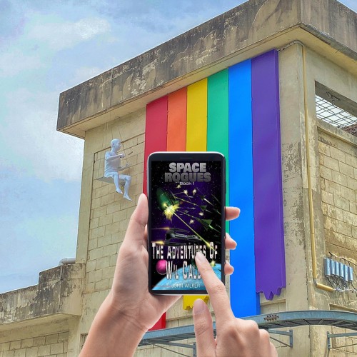 A pair of hands holding an iPhone with the cover of ‘The Adventures of Wil Calder’ on it. In the background a building with an art installation of someone painting rainbow stripes down the side.