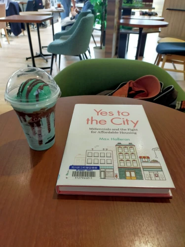 Photo is a round wooden table inside a cafe. The hardcover white library book is in the center with drawings of buildings on it. To the left is a green & brown mint chocolate shake with a scoop of mint chocolate ice cream on top. In the distance you can see many green upholstered chairs and other wooden tables.