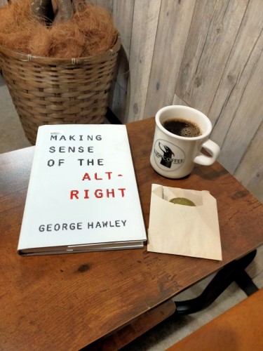 Photo of brown wooden table with the white hardcover book where all the text is black with the exception bring ALT-RIGHT in red  To the right is a green cookie slightly inching out of a light brown paper bag. Above is a light brown mug of black coffee. The mug has a black dog icon along with the coffee house name mentioned in the skeet. The wall is grey wood. In the distance is a dark brown wicker basket