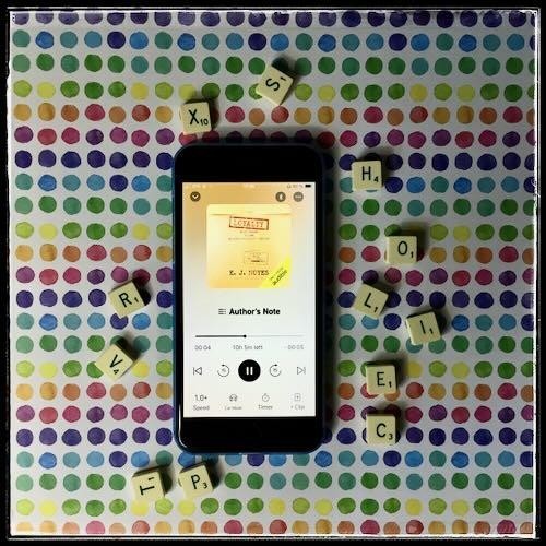 An iPhone with the cover of Loyalty by E. J. Noyes, narrated by Abby Craden, on a white background with numerous multicoloured polka dots. Around the phone are several letters from a game of Scrabble: T - P - V - R - x - S - H - O - L - I - E - C.