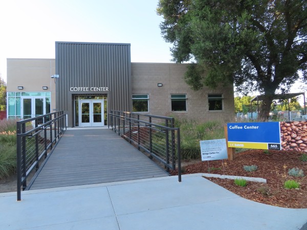 The new one-story UC Davis Coffee Center building, which features a wide ramp bridging a swale between the Arboretum mixed use path and the main entrance to the building. On the right of the ramp is a small grey coroplast sign.