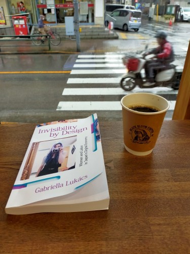 The photo is of a coffee house counter looking out onto a rainy street. The white paperback book is on the brown wooden counter. It features a photo of a young long blue, pink, & black haired Japanese woman in a black tank top & black jeans with a camera covering her nose & mouth. The brown outside/white inside cup of black coffee has a profile of a black and white sloth logo. Outside you can see an intersection. A old blurry Japanese man is heading left on a scooter where another old blurry Japanese man wearing a mask is on a bicycle on the distant sidewalk heading right