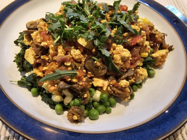 a white and blue plate of food including what might look like egg but is crumbled tofu made yellow by the spice turmeric, as well as mushrooms, tomatoes, onions and peppers on a bed of cooked kale, peas and sweet corn. it is garnished with chopped arugula.