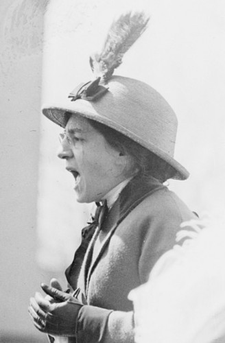 Maud Malone, 1914, in a feathered hat and glasses, overcoat and gloves, giving a speech. By Bain News Service, publisher - File:Maude_Malone_at_women&#039;s_suffrage_meeting,_May_1914.jpg, Public Domain, https://commons.wikimedia.org/w/index.php?curid=81475499