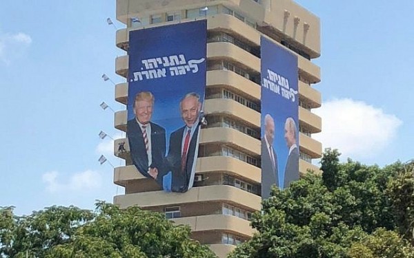 Two massive posters hanging from a tall building. It appears to be about seven stories in height. There are Israeli flags in the corners of every story.

One of the posters feature Donald Trump and Benjamin Netanyahu, close together, smiling, shaking hands, looking outward. Benyahu is wearing a red tie similar to the one's Trump tends to wear.

The other giant poster features Benjamin Netanyahu and Vladimir Putin, looking at provingly at each other.