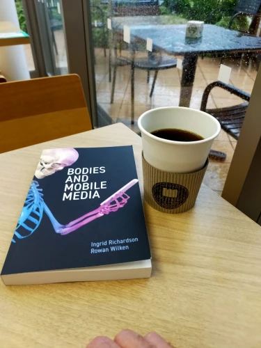 The photo is of inside a cafe and you can see it raining outside via the wetness on a black square metal table outside. Inside the cafe on a light brown wooden table is the black paperback book with a skeleton looking at an extended smartphone. To the right is a white paper coffee of black coffee with a brown coffee jacket with the cafe's logo of a black circle & a loaf of bread in the middle