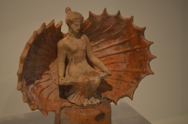 Terracotta figurine of Aphrodite sitting in an open seashell. She has her hair in an updo bun and wears a dress around her legs that she has opened all the way to her knees, revealing her vulva.