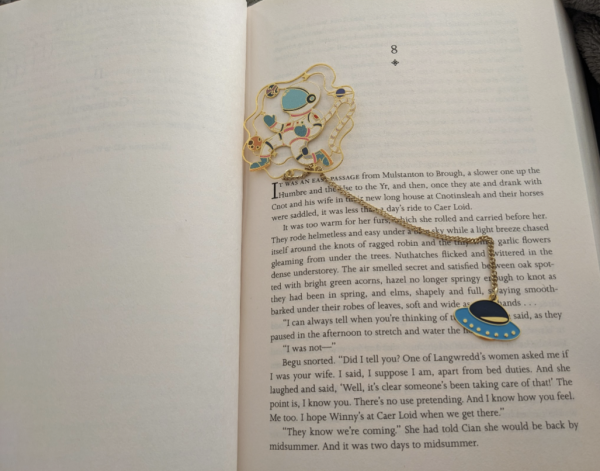 An open book with a blank verso and the beginning of chapter 8 on the recto. Splayed across the page is a metallic bookmark: an astronaut in a white spacesuit with peach, blue, and red accents floats among colorful planets.  A gold chain leads to a blue floating saucer beneath the spacewalker.