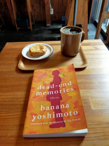 Photo is of inside the cafe. The paperback book is of orange gingko leaves and a carved out red profile of a woman. Above it is a wooden tray on which is a circular white plate with a tan round cookie on it. To its right is a brown mig of black coffee