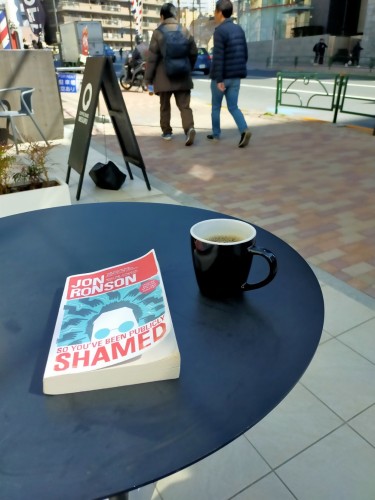 The photo is outside the coffee shop. The metal table is round. The paperback book is on it with an illustration of the eyes & forehead of the author, a white man with short hair and aquamarine glasses. Above his head are a round of hands pointing at him. To the right is a black mug of black coffee. In the distance are 2 Japanese people walking further by. There is a black sandwich board between the people and the table. The sidewalk is red brick