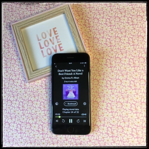 An iPhone with the cover of Don't Want You Like a Best Friend by Emma R. Alban, narrated by Mary Jane Wells & Morag Sims, against a pink flat lay with white flowers. Under the phone, partially hidden, is a small wooden frame with the word "Love" written three times.