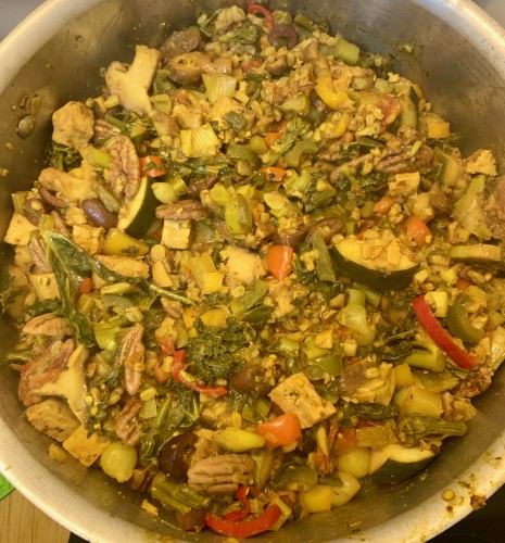 A big pot of stew with an overall yellowish color (from the turmeric). Visible are sliced mushrooms, Kalamata olives, chunks of zucchini, small cubes of tofuk, intact oat grains, broccoli florets, pearl onions, and kale leaves.