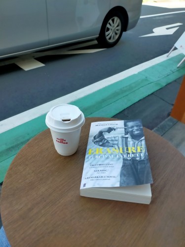 The photo is outside a cafe. On a brown round wooden table is the paperback book with a black and white photo of a young Black boy smiling with a toy gun pointed at his head. He is wearing suspenders holding up jeans  He is also wearing a long sleeve striped dress shirt. The book's title ERASURE is i  uppercase yellow letters. To the right is a white lidded white coffee cup with "mills coffee" in red lowercase letters. Close to the table is a green painted line with white outline and a silver van passing by on the street